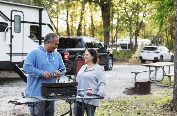 A mature couple, in their 50s, grilling at an RV park, near their camper trailer. 