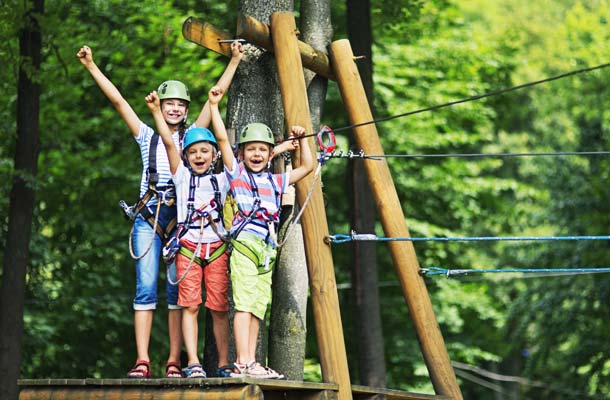 Little girl aged 10 with her brothers aged 7, wearing helmets standing on wooden platform holding zip line in the outdoors ropes course adventure park